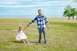Photo of Olger and his daughter holding hands in the midst of green grass on a clear day. He wears a blue flannel and she wears a pink dress, with a matching pink bow in her hair. Both look cheerful.