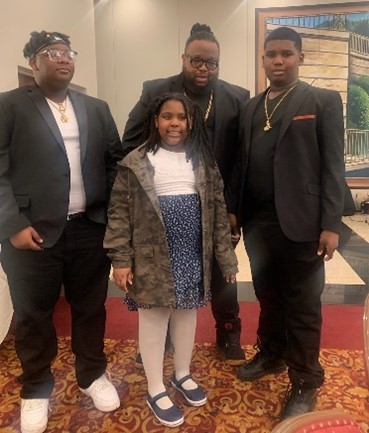 Thai Thergood and his family.  He are all well-dressed, with the boys wearing black suit jackets, and his daughter wearing a skirt and camo jacket.